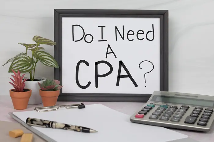 Desk with a framed sign asking 'Do I Need A CPA?' next to a calculator, pen, and notepad, symbolizing the contemplation of hiring a CPA for business financial management.