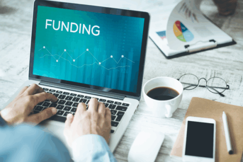 A person using a laptop showing a "FUNDING" screen with financial charts, alongside a coffee cup, glasses, smartphone, and papers on a wooden desk, highlighting business funding tips.